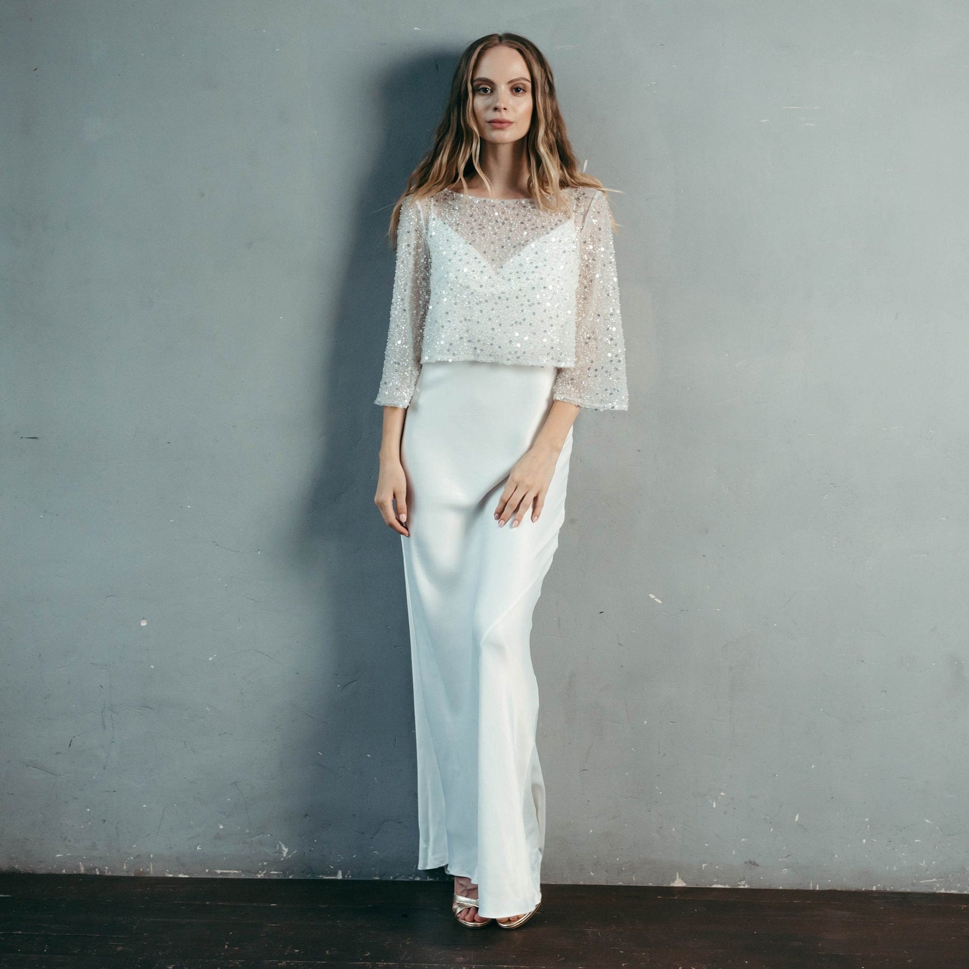 Bride standing against a grey backdrop in a beaded tulle bridal top – lightweight bridal cover-up for wedding or reception.