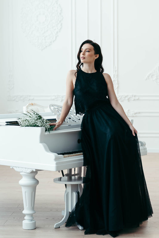 Bride seated in black lace two-piece wedding set with full tulle skirt, posing by white grand piano in a bright studio