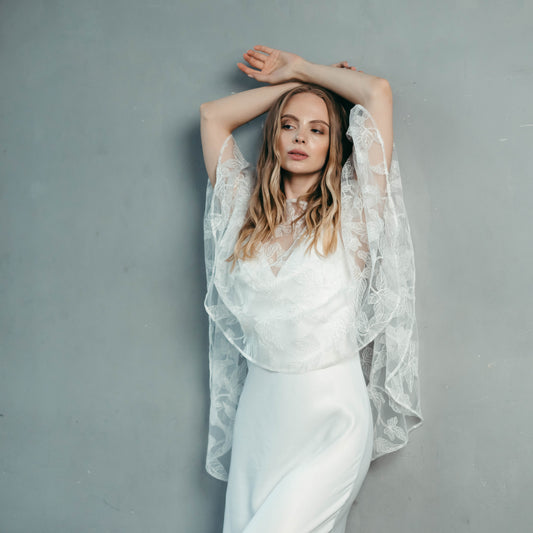 Bride wearing sheer ivy lace bridal capelet over a satin gown, leaning against grey wall with one arm raised