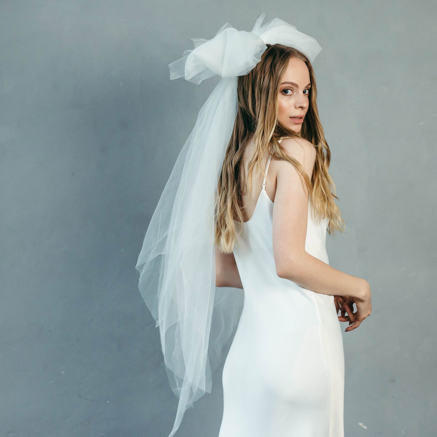 Bride sitting in silky satin slip wedding gown and tulle bow veil, modern romantic styling.