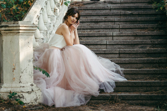 Bride in blush ombré tulle bridal skirt and ivory top sitting on outdoor stone stairs