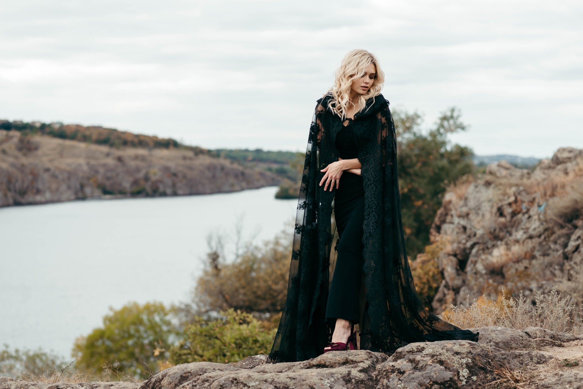 Bride in black lace hooded cape on lakeside cliff for alternative wedding look