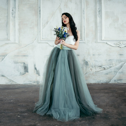 Bride wearing ombre tulle wedding skirt in sage and moss green, holding a bouquet against a textured white wall – bridal separates for outdoor weddings.