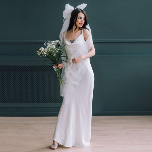 Bride wearing silky satin slip wedding dress with oversized bridal bow veil and bouquet.