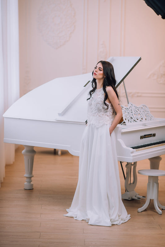 Bride in ivory lace bridal top and flowing crepe skirt standing beside a white grand piano