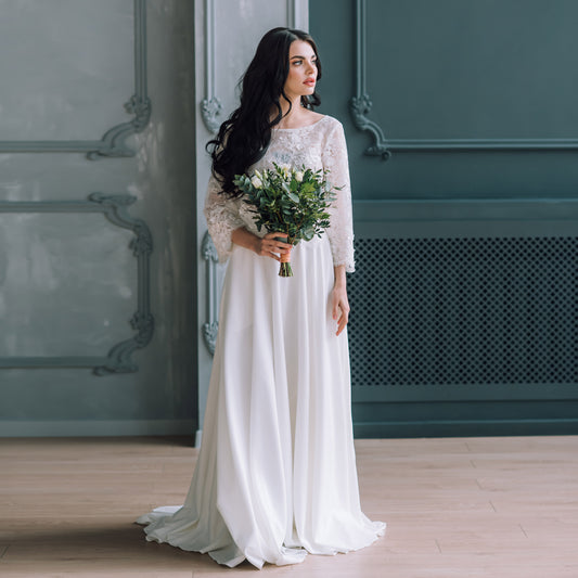 Model wearing long white bridal skirt with fitted waistband styled with long sleeve lace top, holding bouquet.