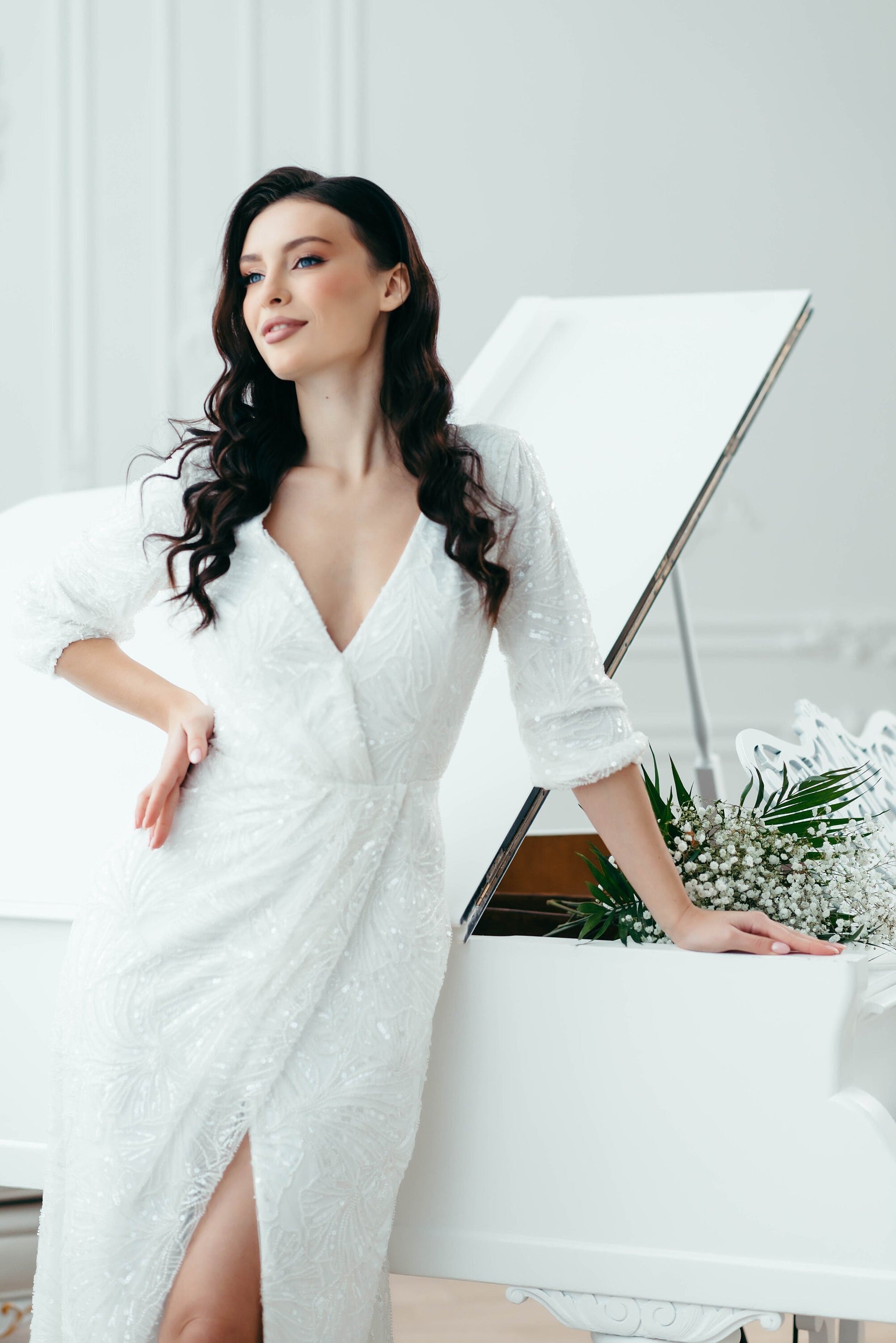 Bride sitting at white piano wearing floor-length beaded wedding dress with high slit