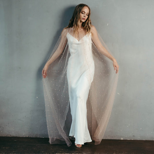 Bride wearing blush pink pearl tulle cape over ivory slip dress for romantic wedding look.