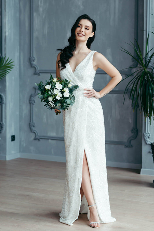 Bride wearing ivory beaded guipure lace wedding dress with high front slit, sleeveless design, holding bouquet – elegant modern bridal gown.