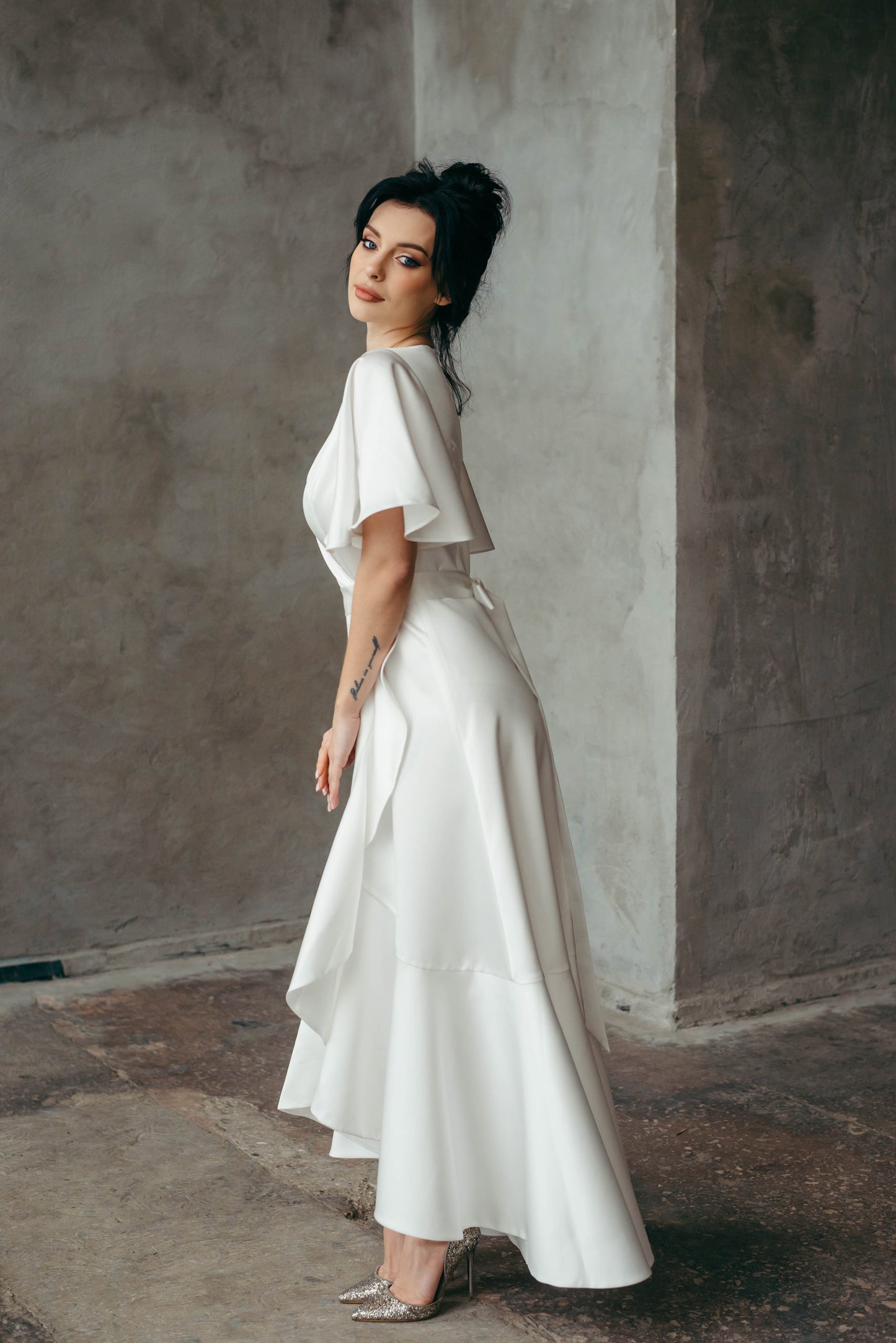 Woman in a white dress standing against a textured wall. Back view of a satin bridal gown, highlighting boho silhouette.