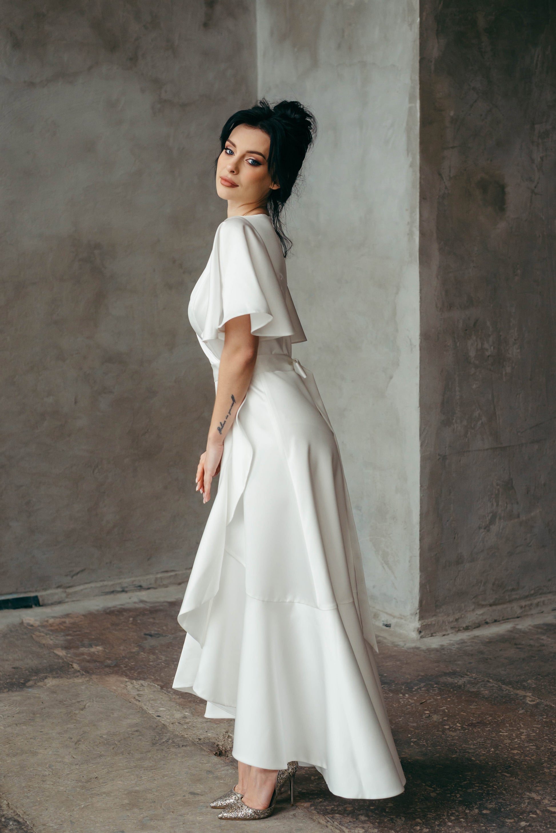 Woman in a white dress standing against a textured wall. Back view of a satin bridal gown, highlighting boho silhouette.