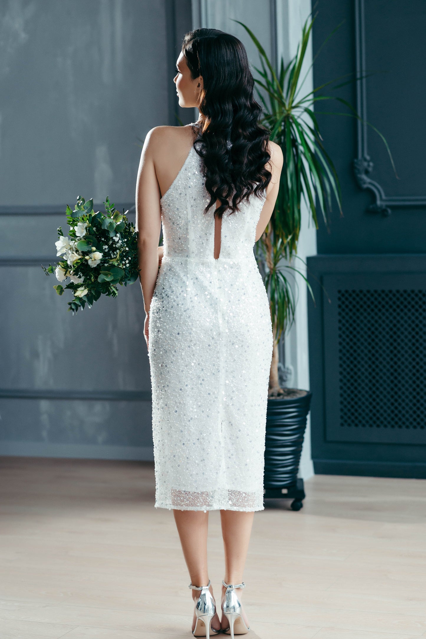 Woman in a white lace midi wedding dress holding flowers in a room with a plant and dark wall.