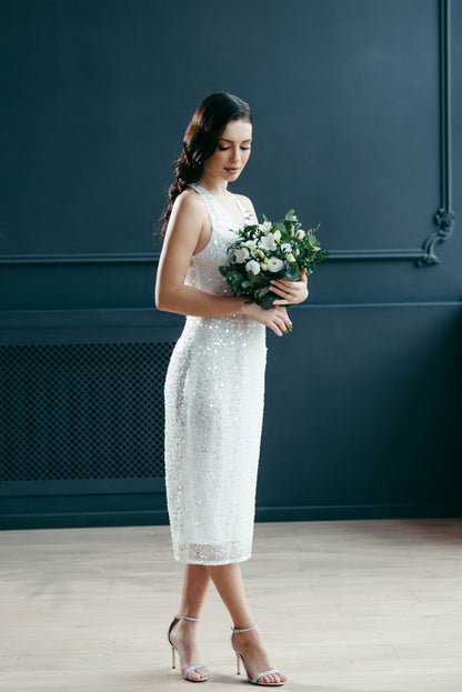 Bride in a midi beaded wedding dress holding a bouquet against a dark blue wall.