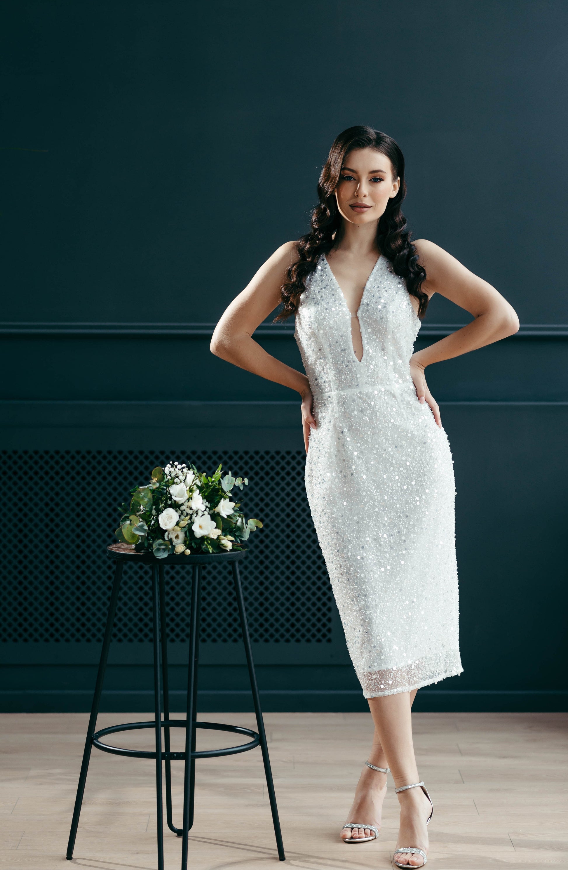 Bride in a white beaded elopement dress standing next to a floral arrangement on a stool against a dark blue wall.