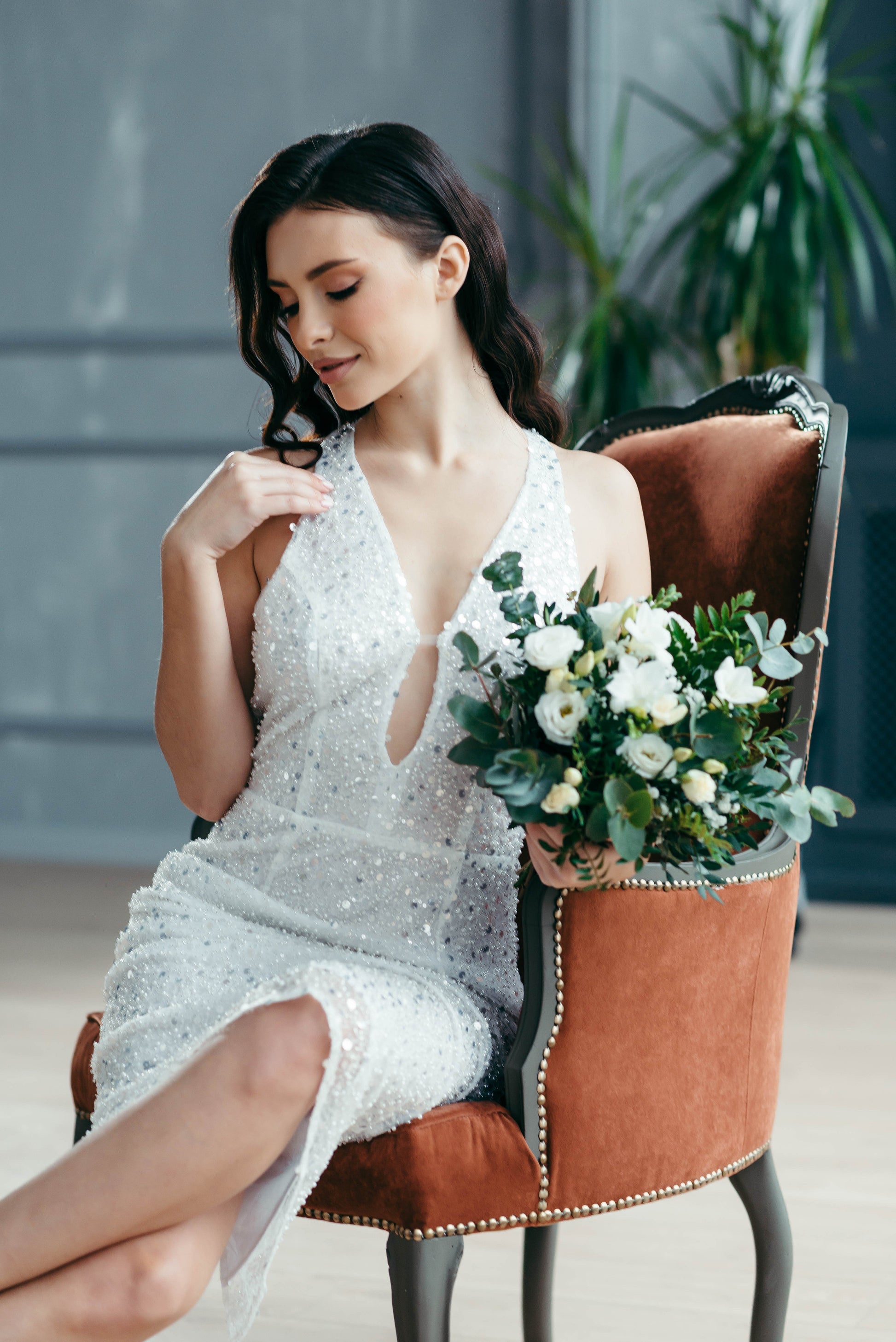 Woman in a short beaded wedding dress holding a bouquet of flowers, sitting on a brown chair.