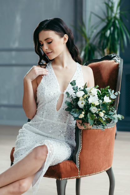 Woman in a short beaded wedding dress holding a bouquet of flowers, sitting on a brown chair.