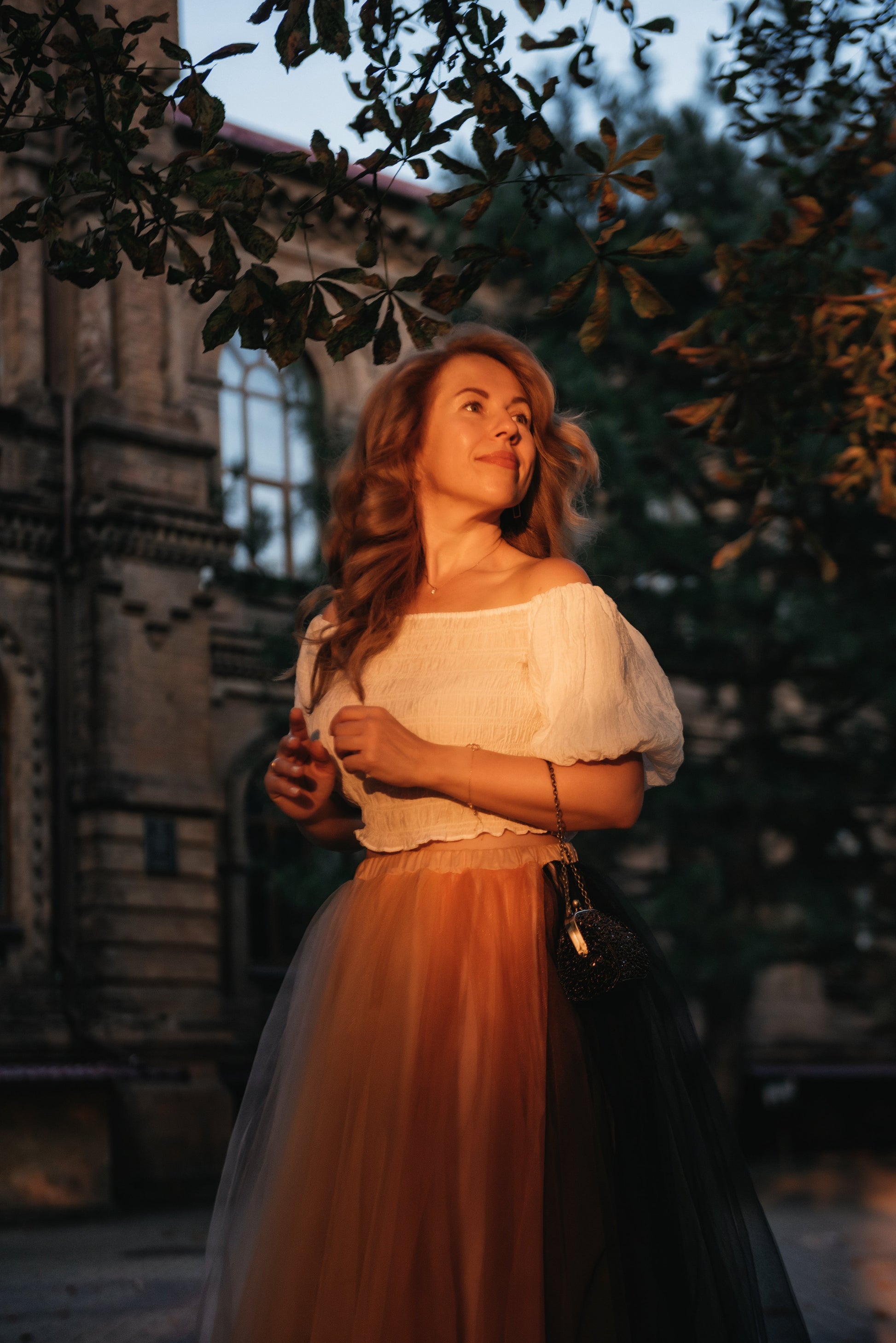 Woman in a white blouse and multicoloured bridal skirt standing outdoors with trees and building in the background