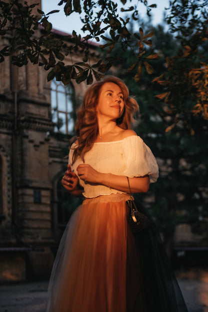 Woman in a white blouse and multicoloured bridal skirt standing outdoors with trees and building in the background