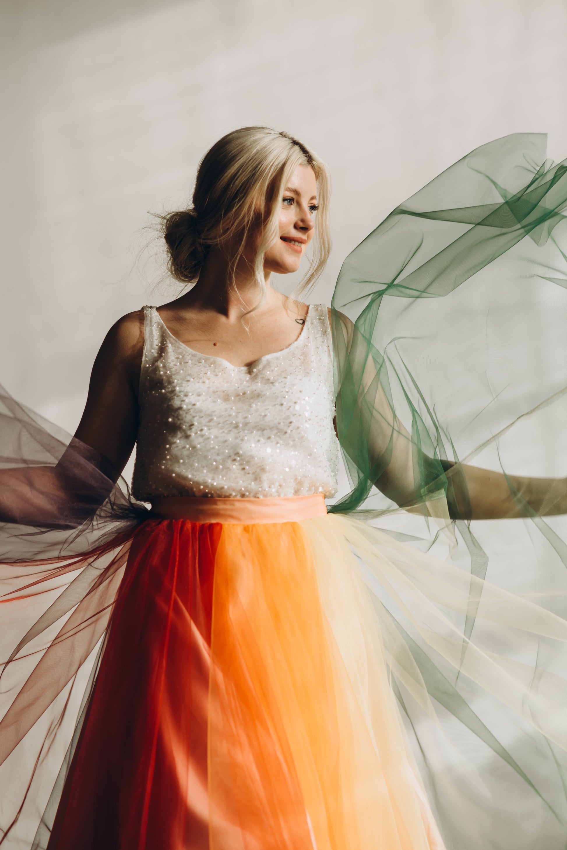 Bride in a white beaded top and rainbow wedding skirt with flowing fabric against a neutral background