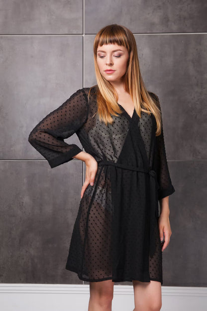 Woman wearing a black sexy sheer dressing gown with sheer panels against a gray tiled wall.