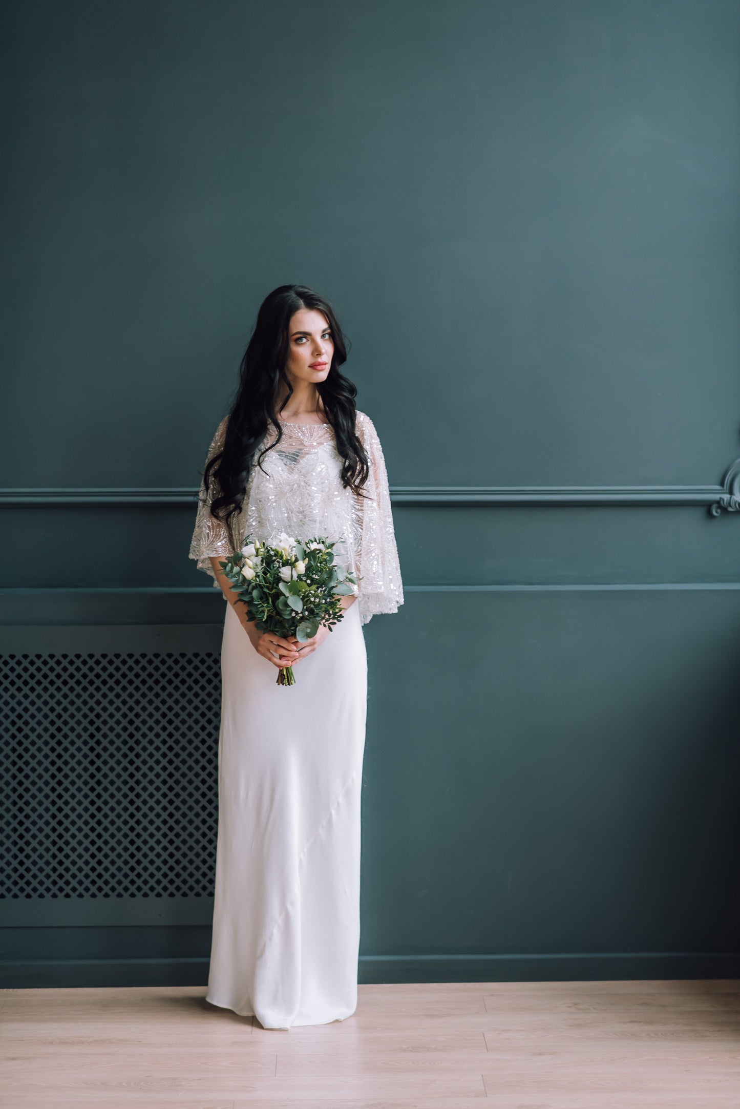 Bride holding a bouquet against a dark green wall wearing a simple dress and a sparkle bridal cover up