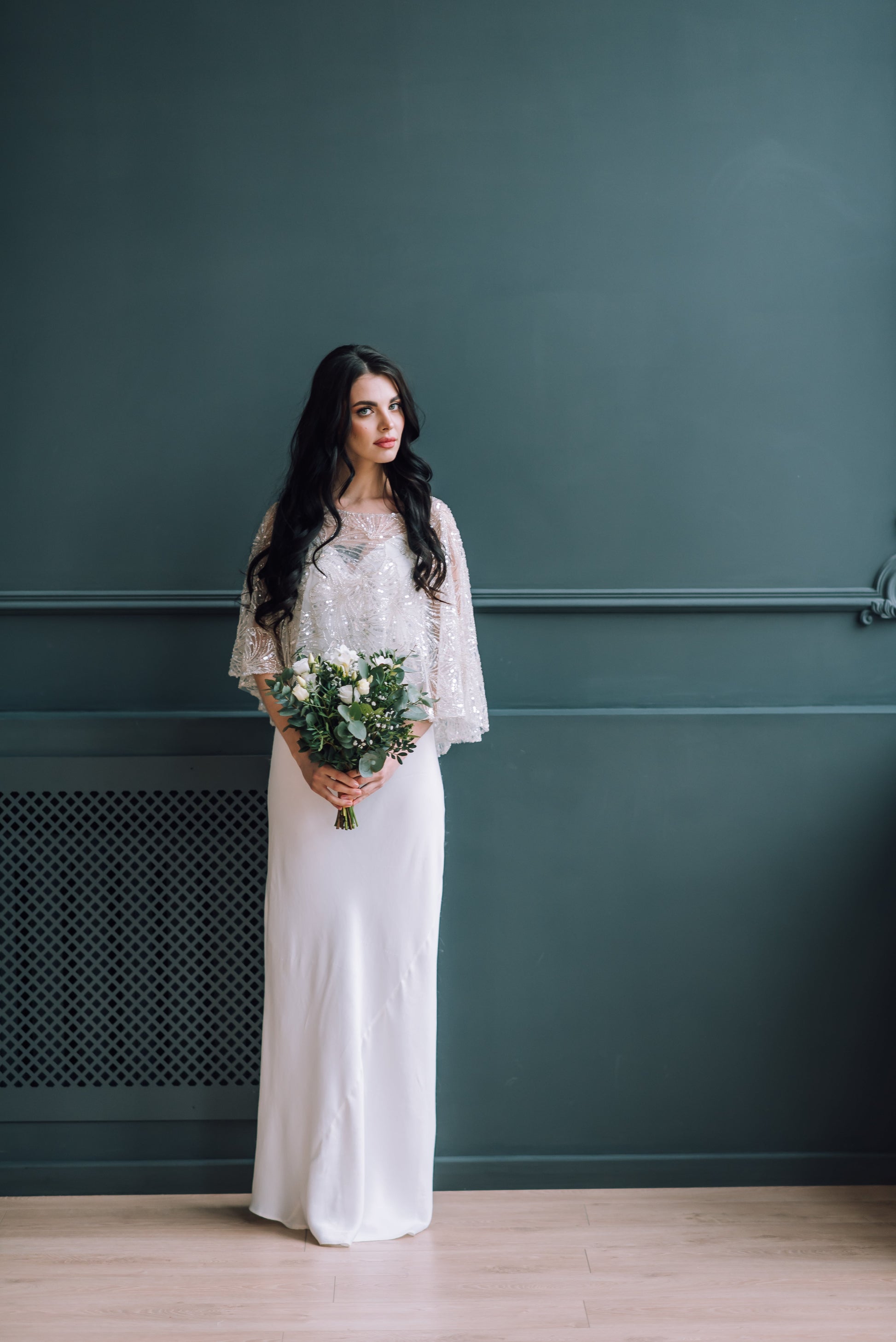 Bride holding a bouquet against a dark green wall wearing a simple dress and a sparkle bridal cover up