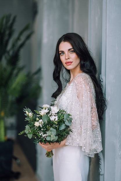 Bride in a white dress and handmade sequined and beaded capelet holding a bouquet of flowers indoors